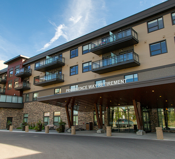 Prominence Way Retirement building exterior with balconies above a covered entrance, blue sky behind.