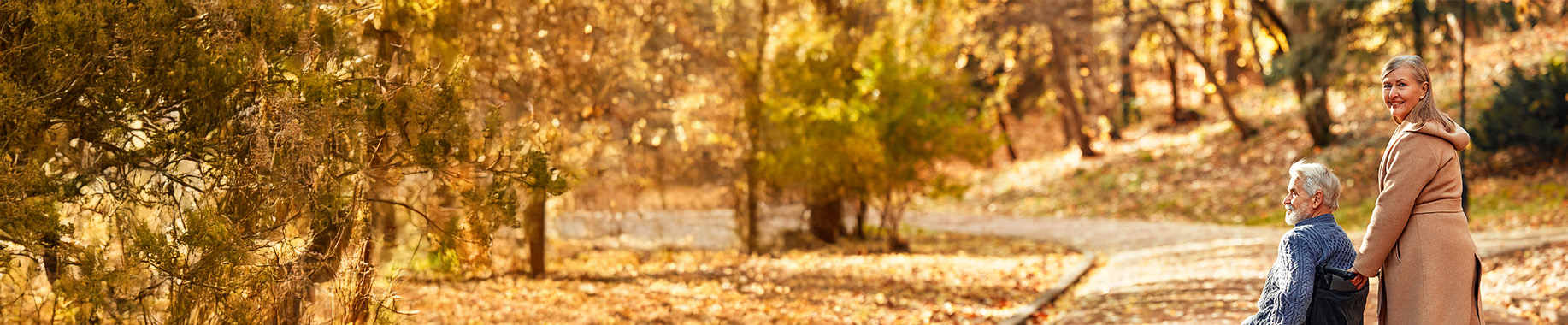 Woman pushing a man in a wheelchair on a path, autumn trees in front and behind them.