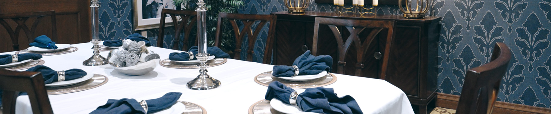 Formal dining table set with navy napkins and silver candlesticks, patterned wallpaper behind.