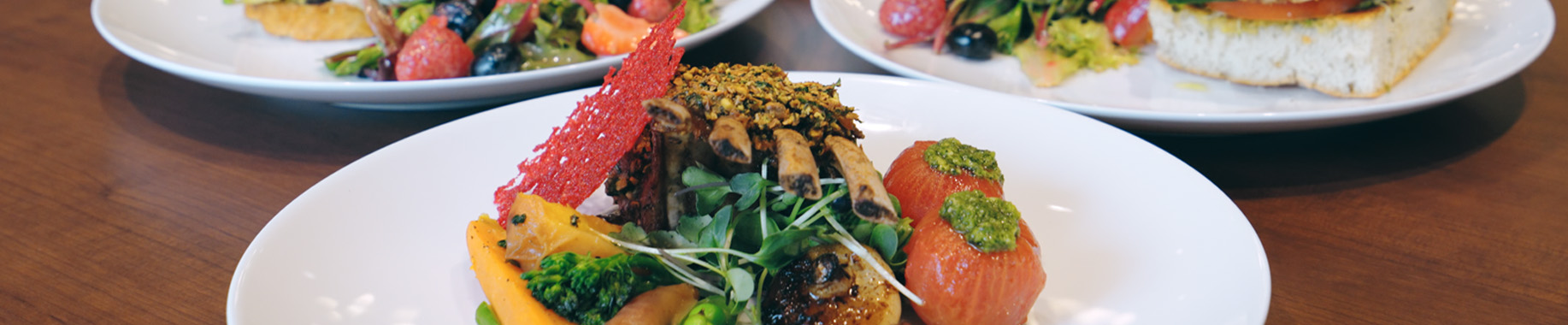 Rack of lamb with roasted vegetables in front, salad and sandwich plates behind on a wood table.