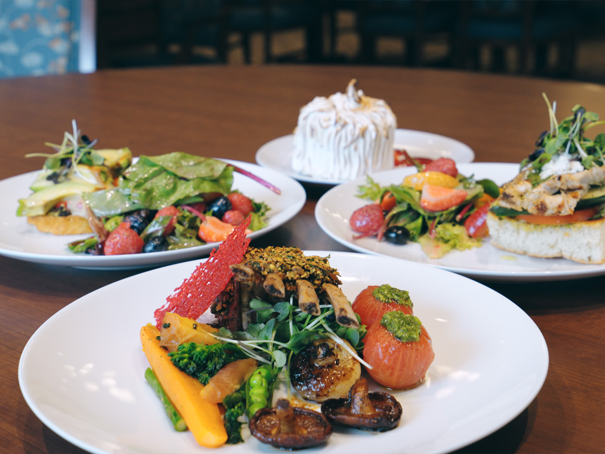 Rack of lamb with vegetables in front, salads, and cake plated on a wood table.