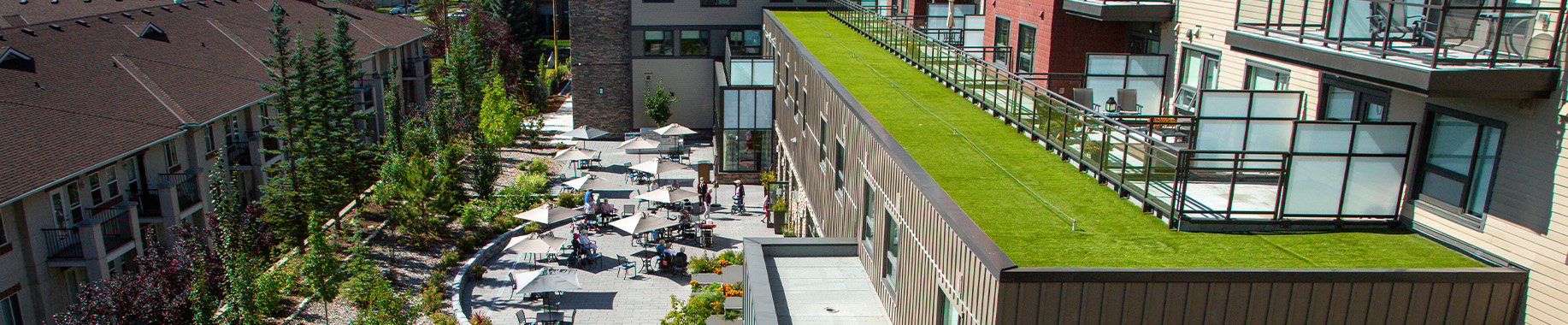 Aerial view of a courtyard patio with umbrella tables, green rooftop terrace beside residential buildings.