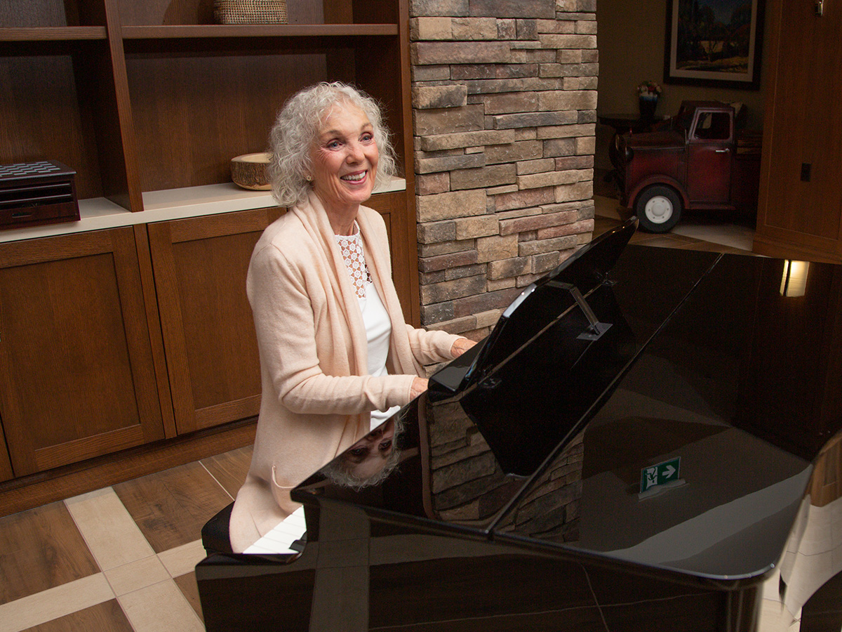 Smiling older woman playing a black grand piano, stone fireplace visible behind her.