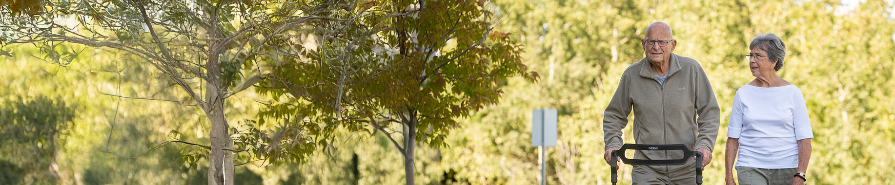 Older couple walking on a path with a walker, trees in front and behind them.