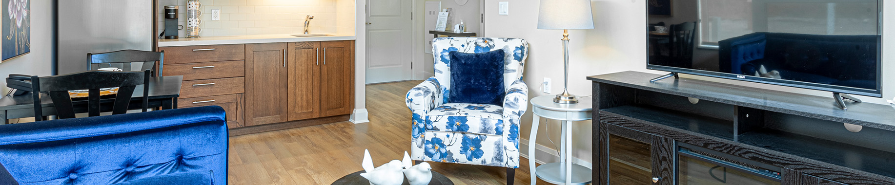 Living room with a blue sofa in front, floral armchair beside a TV console, kitchen behind.