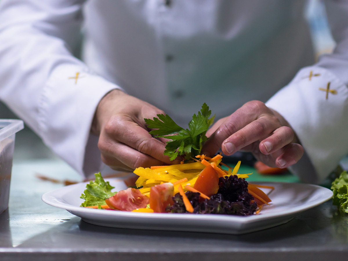 Chef plating a colourful vegetable salad with garnish on a white plate in a kitchen.