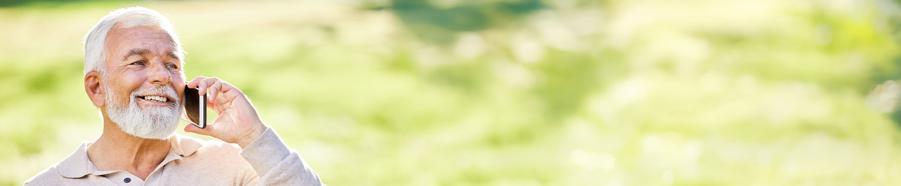 Smiling older man talking on a phone outdoors with green foliage blurred behind him.