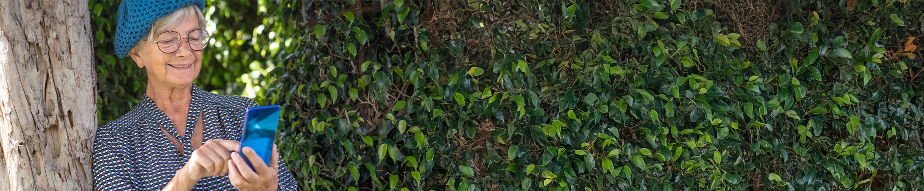 Older woman wearing a blue knit hat and glasses using a smartphone beside a leafy hedge.