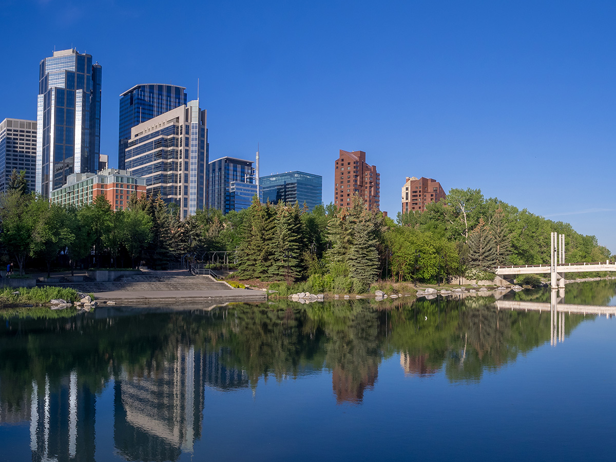 Calgary skyline behind trees and a footbridge reflected in calm water under a clear blue sky.